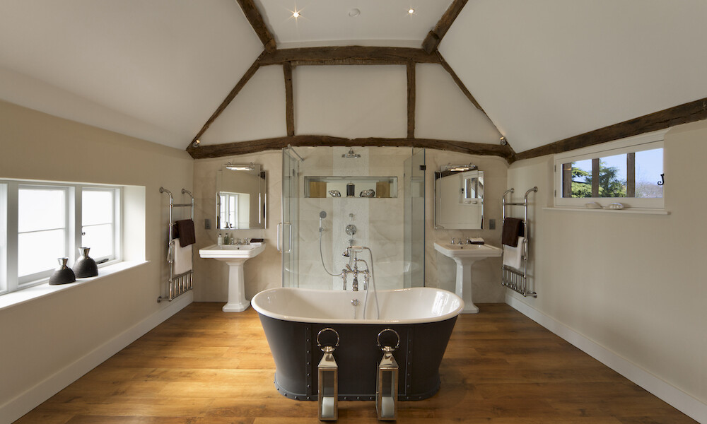 "a beautifully prepared bathroom in a 500 year old farm building that has been completely restored as a new home. Old weathered oak beams support the roof and ceiling whilst in the centre of the new oak flooring sits a traditional styled large bath with old fashioned taps and shower. In the background, a more contemporary shower unit and screens can be seen, either side of which are two large traditionally styled basins and taps. Note the heavy old-styled towel rails. Two lanterns sit in front of the bath...Looking for a Bathroom image Then please see my other Bathrooms and related inages by clicking on the Lightbox link below...A>AA>A"