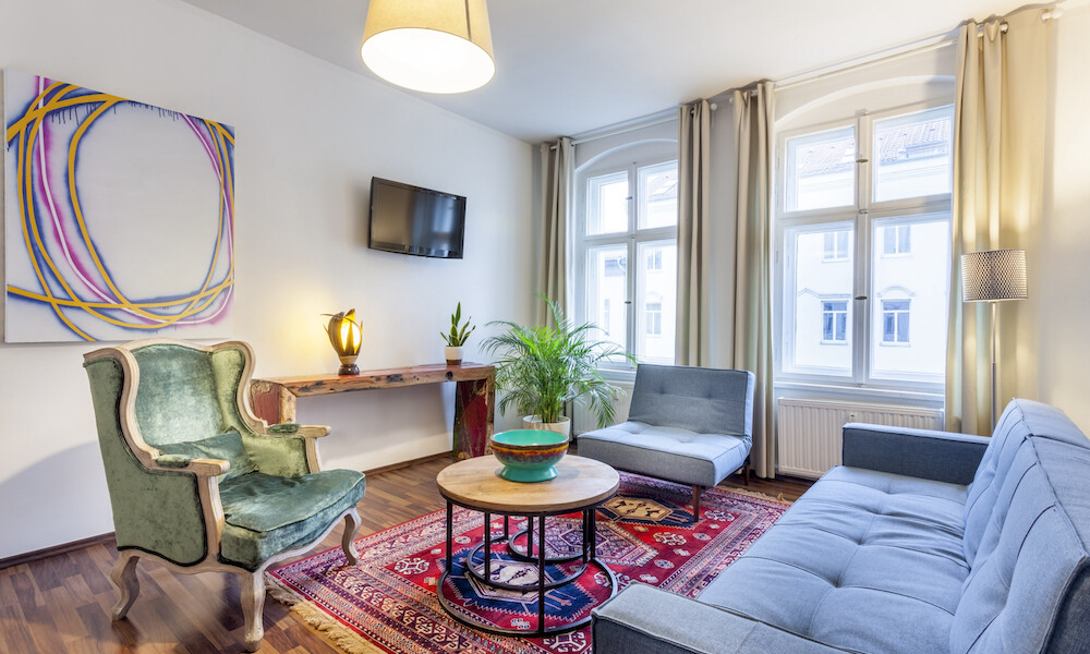 Classic styled interior of a living room with a Danish designer couch, coffee table and Persian carpet. There is also a sideboard above with is a wall mounted flat screen television. On the wall hangs artwork in the forma of an acrylic painting. The floor is laminate wood.